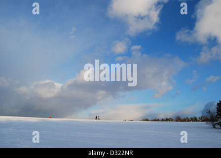 Einen dramatischen Verlauf der dunklen und hellen blauen Himmel und Wolken mit Blick auf eine große Decke von Schnee auf einem Feld mit Menschen am Horizont Stockfoto