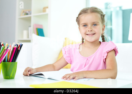 Niedliche kleine Mädchen vom Tisch mit Buntstiften und offene Copybook vor ihr sitzt Stockfoto