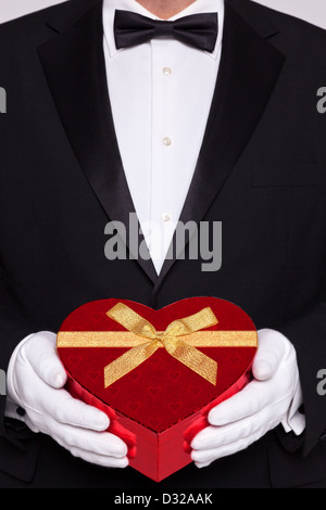 Mann trägt schwarze Krawatte und weiße Handschuhe halten eine rote herzförmige Schachtel Pralinen. Stockfoto