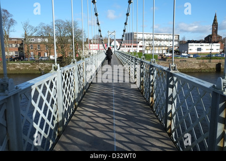 Eine Frau in roter Schal Spaziergänge auf der anderen Seite des Flusses Nith Suspension Bridge, Dumfries, Schottland. Ganz rechts ist St Michaels, Begräbnisstätte von Robert Burns Stockfoto