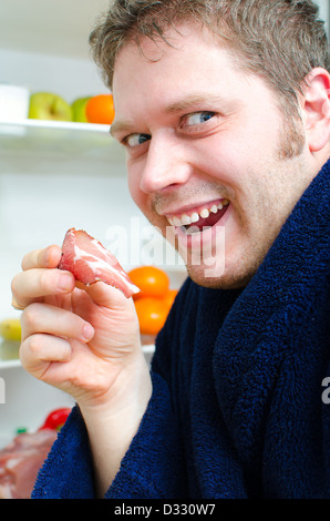 Gut aussehender Mann Essen Schinken Scheibe in der Nähe von offenen Kühlschrank Stockfoto