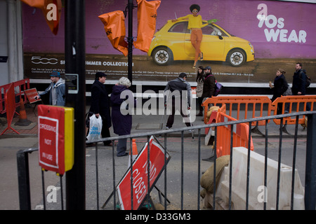 Fußgänger laufen unter einem breiten Plakatwerbung der Fiat 500 mit einem 70er Modethema. Ein Mann auf Krücken geht unbeholfen auf der anderen Straßenseite und auf die weit Bürgersteig unter dem Bild des Modells und das gelbe Auto Fiat. Baustellen werden auf dieser Kreuzung, Teil der Regeneration des nahe gelegenen Hauptstrecke Bahnhof London Bridge durchgeführt. Die Straße ist gesperrt und die Überfahrt nicht verwenden, auf der Südseite der Themse im Stadtteil Southwark. Stockfoto