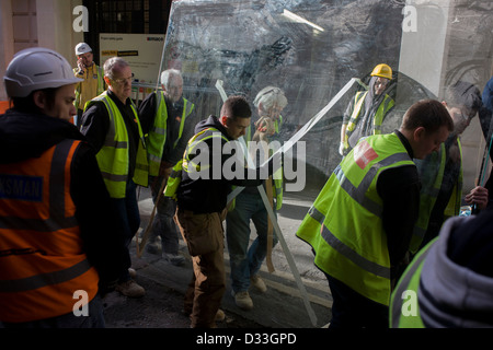 Ein Team von Arbeiter tragen-Sichtbarkeit Wappenröcke mit dem Namen des Bau Firma Barnwood auf der Rückseite, kämpfen, um eine sehr schwere Plattenglas-Fenster durch eine Stadt der London Street zu hieven. Unter einem kein Eintrag Verkehrsschild vorbei, gehen die Männer auf dem Bürgersteig gegenüber einem geparkten LKW, der seine nächste Ladung im Herzen des Finanzviertels der Hauptstadt bekannt als die quadratische Meile nach seinen kreisenden römische Stadtmauer erwartet. Stockfoto