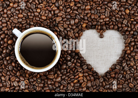 Herzform aus Kaffeebohnen mit einer Tasse Kaffee auf Leinenstoff hergestellt. Stockfoto