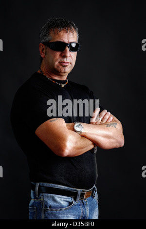 Düsterer harter Kerl mit Tatoos und schwarze Sonnenbrillen, Arme verschränkt, Blick in die Kamera, Studio über Schwarz erschossen. Stockfoto