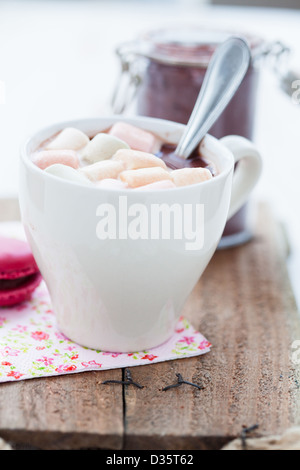 Nahaufnahme der Tasse heiße Schokolade mit farbigen Marshmallows ruht auf Holztisch mit Glas Kakaopulver im Hintergrund Stockfoto