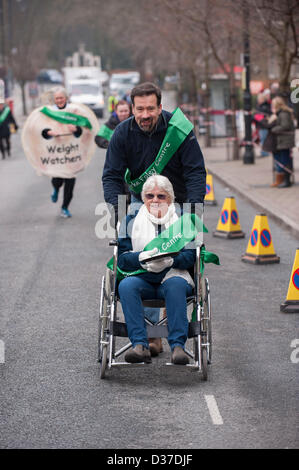 Wettbewerber (Mann, alte Dame im Rollstuhl), die sich an traditionellen Ilkley Rotary Pancake Race - The Grove, Ilkley, West Yorkshire, UK. Stockfoto