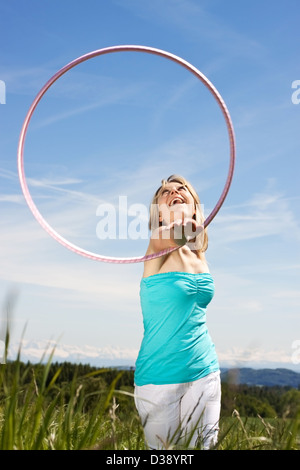 gerne hübsche Frau, stehend auf einer Wiese und spielt mit Hula Hoops. Im Hintergrund Berge Stockfoto