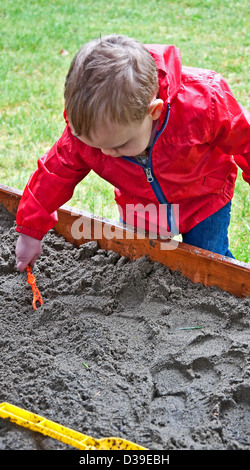 Dieser 18 Monate altes Kleinkind junge spielt in einer Sandbox und Graben im Sand an einem regnerischen Tag. Er trägt seine roten Regenmantel. Stockfoto