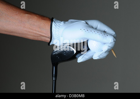 Nahaufnahme einer Golfer Hand hält einen Schlägerkopf, Golfball und Abschlag über eine leichte ot dunkelgrauen Hintergrund. Stockfoto