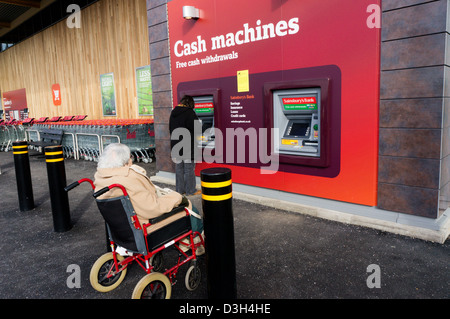 Eine ältere Dame wartet in einem Rollstuhl, während ihre pflegeperson oder Assistant zieht Geld aus einem Geldautomaten in einem Supermarkt. Stockfoto