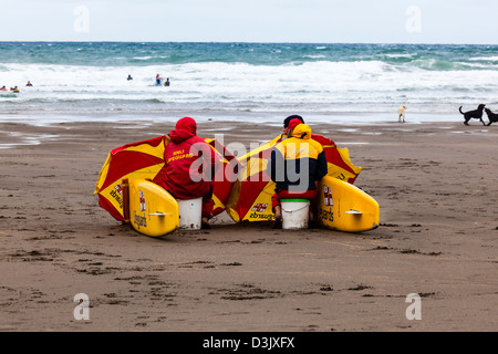 RNLI Rettungsschwimmer verwenden, Surfbretter und Sonnenschirme um Schutz vor dem kalten Wind auf der Black Rock Beach, Widemouth Bay, Nr. Bude, Cornwall, UK Stockfoto