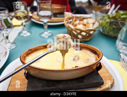 Leckeren Fondue Käse geschmolzen über Brot auf eine Gabel mit einem Hintergrund von einem vollen Tisch von einem freundlichen Mittagessen mit Brot und Wein. Stockfoto