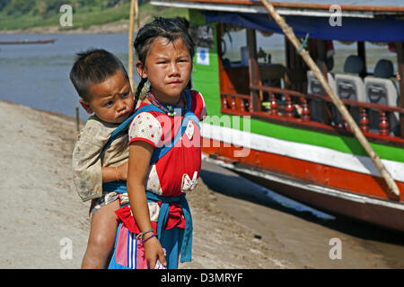 Mädchen mit Baby auf Rücken vor hölzernen Longtail-Boot entlang des Mekong in Laos, Südostasien Stockfoto
