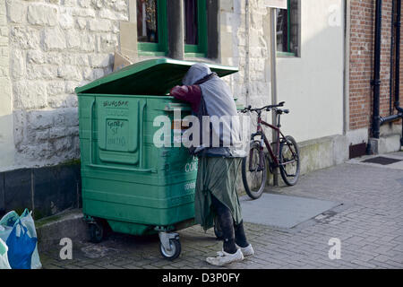 Ein Stadtstreicherin Bettler in Lumpen gekleidet fallend in eine grüne Mülltonne Stockfoto