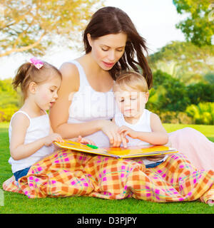 Bild der süße junge Frau mit zwei kleinen Kindern lesen Buch im Freien, Bruder und Schwester mit Mutter lernen im park Stockfoto