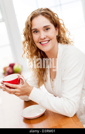 Porträt von lächelnden Frau halten rote Tasse Kaffee am Tisch in der Küche zu Hause Fenster Stockfoto