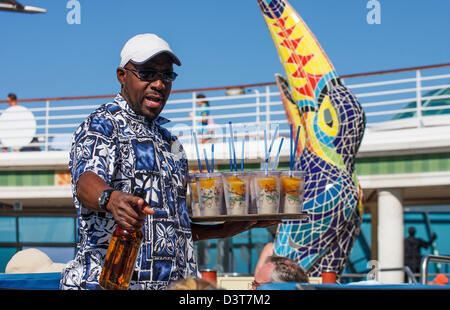 Ein Kellner in einem tropischen Hemd mit einem Tablett an Getränken darauf mit einer Flasche Rum auf dem Deck eines Kreuzfahrtschiffes Stockfoto