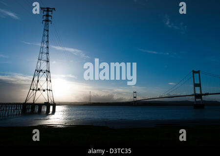 Pylone mit elektrischen Leitungen nach Wales über den Fluss Severn mit Sonne im Wasser und erste Severn-Brücke in Ferne reflektiert. Stockfoto