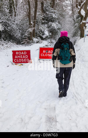 Eine Frau, gekleidet für den Winter Spaziergänge im Schnee vorbei "vorne geschlossen" Verkehrszeichen auf einer Schnee bedeckten Landstrasse, Surrey, England. Stockfoto