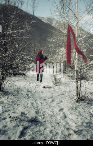 eine junge Frau in einem roten Mantel läuft durch den winterlichen Wald und verlor ihren roten Schal Stockfoto