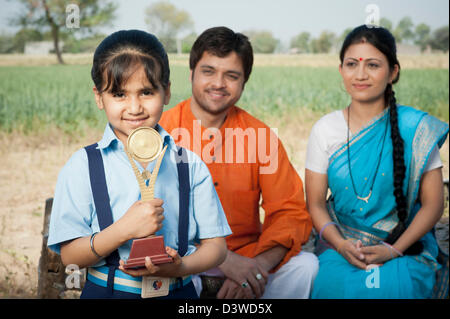 Schulmädchen halten eine Trophäe mit ihren Eltern im Hintergrund, Kapur, Haryana, Indien Stockfoto