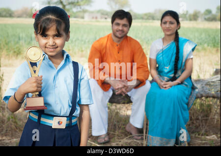 Schulmädchen halten eine Trophäe mit ihren Eltern im Hintergrund, Kapur, Haryana, Indien Stockfoto