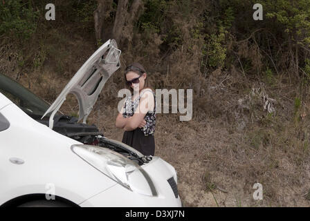 Junge Frau mit Arm gekreuzt schaut ängstlich ihr kaputt Auto mit der Motorhaube auf. Stockfoto