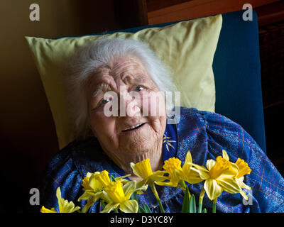 ÄLTERE DAME BLUMEN PFLEGE zufrieden glücklich lächelnd unabhängige 99 Jahre alte ältere Dame sitzt im Zimmer mit einer Besucherpräsentation von Narzissen Blumen Stockfoto