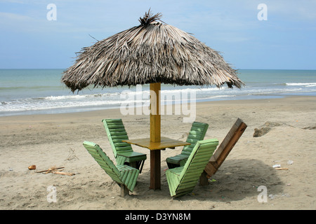 Ein Holztisch mit Stühlen unter einem reetgedeckten Dach auf einem überdachten Sandstrand am Pazifischen Ozean in Tonsupa, Ecuador Stockfoto