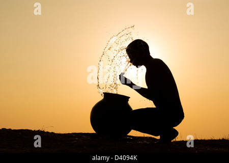 Indischer Mann Spritzwasser über sich selbst aus einem Wassertopf bei Sonnenuntergang. Silhouette. Indien Stockfoto