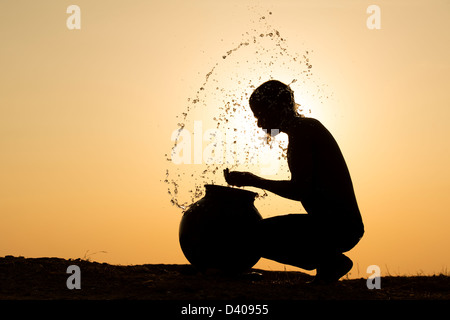 Indischer Mann Spritzwasser über sich selbst aus einem Wassertopf bei Sonnenuntergang. Silhouette. Indien Stockfoto