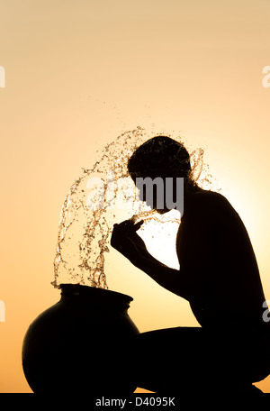 Indischer Mann Spritzwasser über sich selbst aus einem Wassertopf bei Sonnenuntergang. Silhouette. Indien Stockfoto