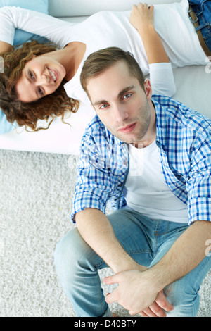 Junger Mann auf dem Boden sitzen und Blick in die Kamera mit seiner Freundin auf Sofa auf Hintergrund Stockfoto