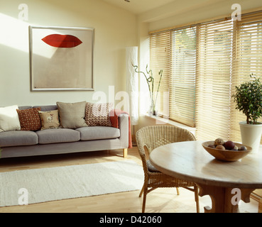 Gemälde an der Wand über dem grauen Sofa mit gemusterten Kissen in modernen Wohn- und Esszimmer mit Lattenrost aus Holz Jalousien Stockfoto