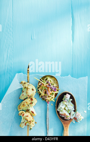 Draufsicht auf gegrillter Blumenkohl auf Screwer und Löffel gefüllt mit Salat auf Wachspapier auf blauen Holztisch im Studio Stockfoto