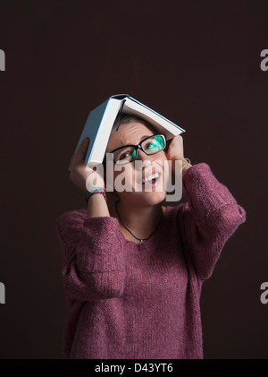 Porträt von Teenager-Mädchen tragen Brillen, halten aufgeschlagenes Buch über den Kopf mit ängstlichen Ausdruck auf schwarzen Hintergrund Stockfoto