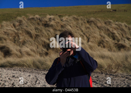 Eine Nahaufnahme von einem Fotografen hält eine Kamera an einem Strand mit grasbewachsenen Dünen und Felder hinter Stockfoto