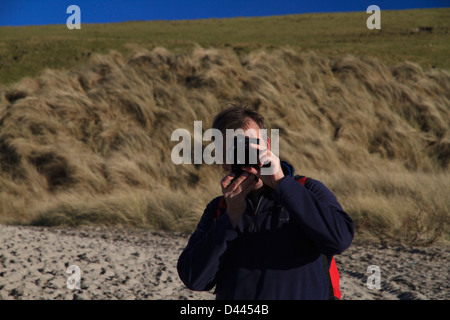 Eine Nahaufnahme von einem Fotografen hält eine Kamera an einem Strand mit grasbewachsenen Dünen und Felder hinter Stockfoto