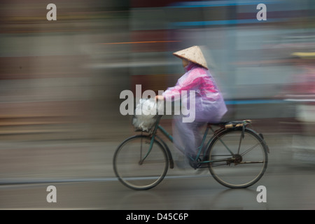 Frau in einem konischen Hut mit dem Fahrrad im Regen, Hanoi, Vietnam Stockfoto