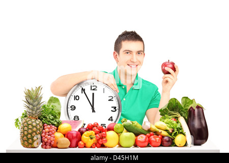 Junger Mann hält eine Wanduhr und Apfel, auf einem Tisch voller Obst und Gemüse, isoliert auf weißem Hintergrund Stockfoto