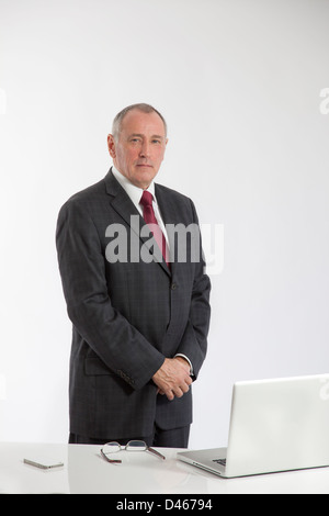 Portrait über eine ältere männliche Büroangestellte hinter einen weißen Schreibtisch mit einem Laptop geöffnet, Gläser und Iphone auf dem Schreibtisch stehen. Stockfoto