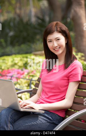 Junge Chinesin mit Laptop, während, entspannend auf Parkbank Stockfoto