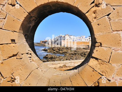 Blick durch ein Bullauge in einer Ziegelmauer auf die felsige Küste vor der befestigten Stadt Essaouira, Marokko Stockfoto