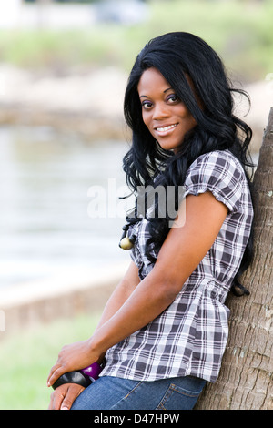 Lässig gekleidet glückliche junge afrikanische amerikanische schwarze Frau lehnt sich gegen einen Baum lächelnd. Stockfoto