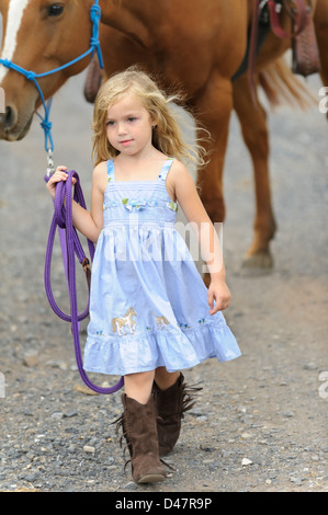 Kleine blonde Mädchen selbstbewusst führt ihr großes Pferd auf einen Feldweg während des Tragens Stiefel und ein blaues Kleid. Stockfoto