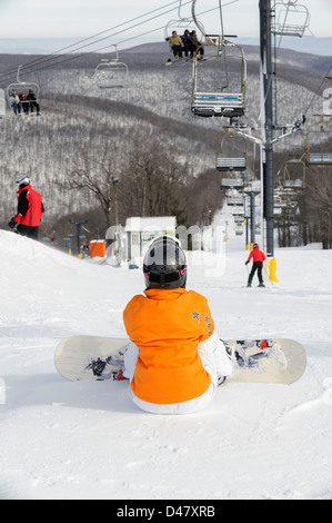 Frau mit Snowboard sitzen und warten am oberen Rand einer Skipiste mit Skilift und Gebirgshintergrund. Stockfoto