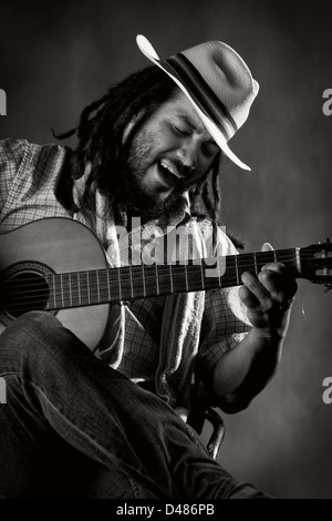 Ein schwarz-weißes Porträt eines Mannes mit langen Dreadlocks und Bart, der einen hellen Fedora-Hut mit gestreiftem Band und einem karierten Hemd trägt. Er sitzt und spielt mit seiner Akustikgitarre Stockfoto