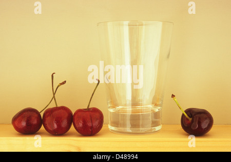 Augenhöhe Blick auf drei dunklen roten Kirschen mit Stielen aufgereiht auf Holzregal mit Glas und vierten Kirsche Stockfoto