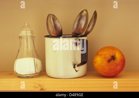 Augenhöhe Blick auf schmalen Holzregal mit halbvollen durchsichtigen Kunststoff Salz Menage Emaille Becher mit drei Teelöffel und Apfel Stockfoto